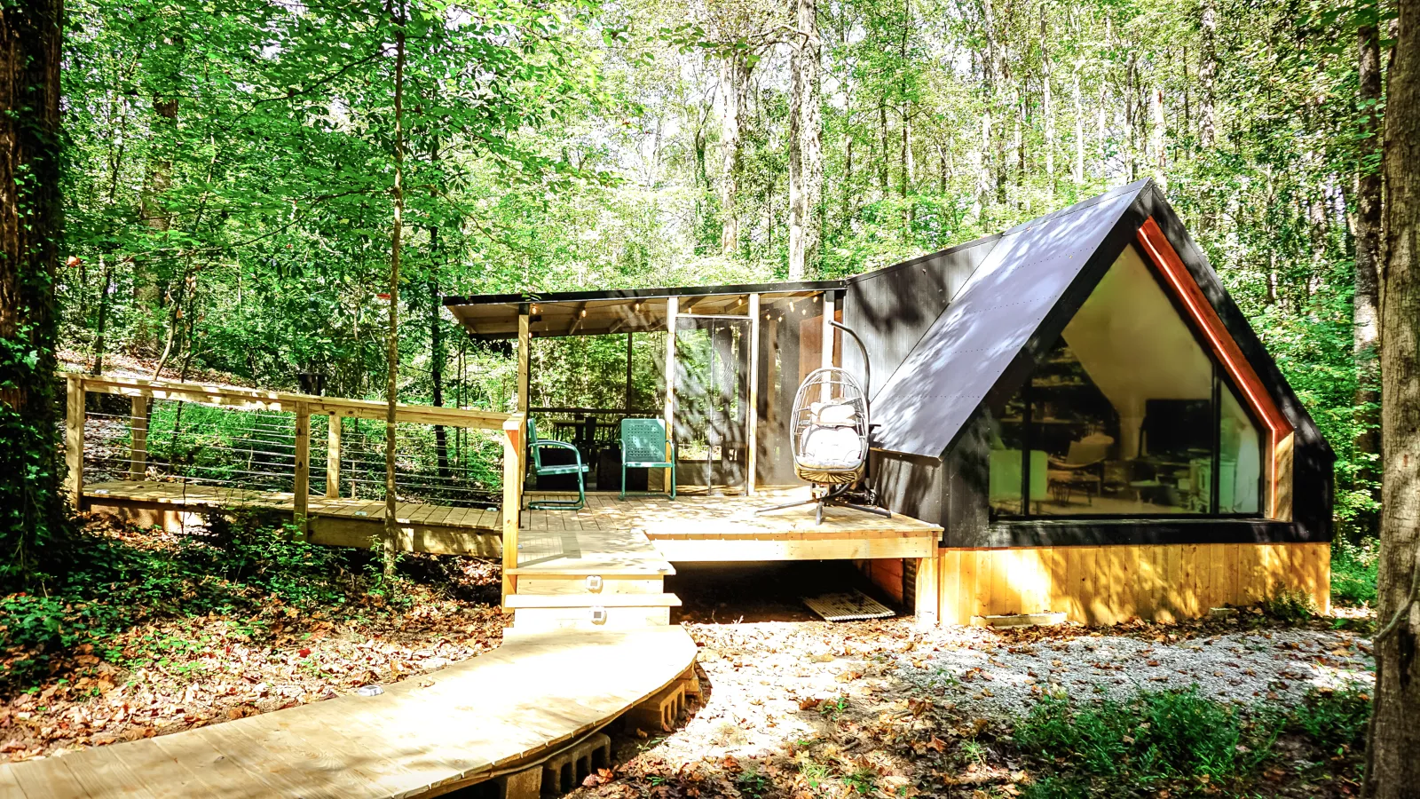 The A-frame cabin and hanging egg chair reached by a curved boardwalk through sunlit forest